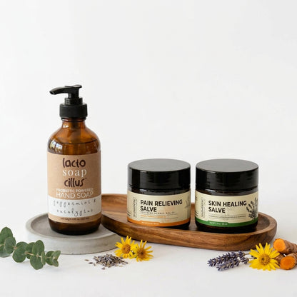 Amber pump bottle of probiotic hand soap next to two dark glass balm jars on a wooden tray, with eucalyptus, lavender, yellow flowers and turmeric root