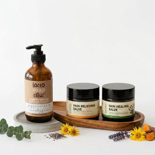 Amber pump bottle of probiotic hand soap next to two dark glass balm jars on a wooden tray, with eucalyptus, lavender, yellow flowers and turmeric root