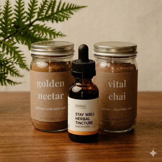 Three packaged wellness items arranged on a wooden surface: two glass mason jars labeled golden nectar and vital chai, and one amber dropper bottle labeled stay well herbal tincture. A green fern plant is visible in the background against a light neutral wall.