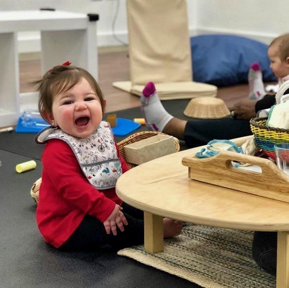 Smiling baby in red shirt sits by sensory play table with Montessori toys in classroom setting