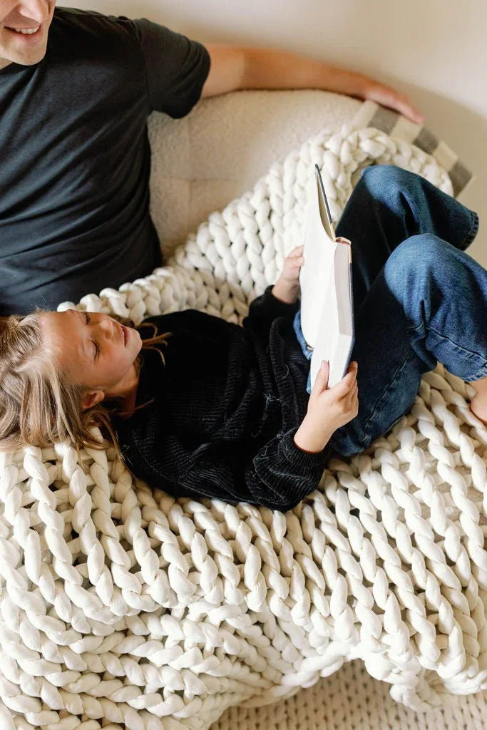 Child reading a book while lying on a chunky knit sensory blanket next to an adult