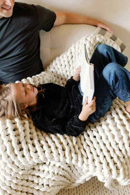 Child reading a book while lying on a chunky knit sensory blanket next to an adult