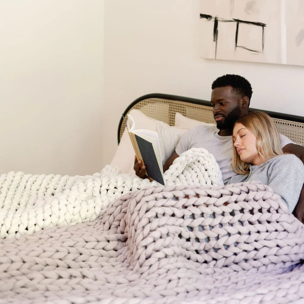 Couple relaxing in bed under chunky knit sensory blanket, reading together in cozy bedroom.
