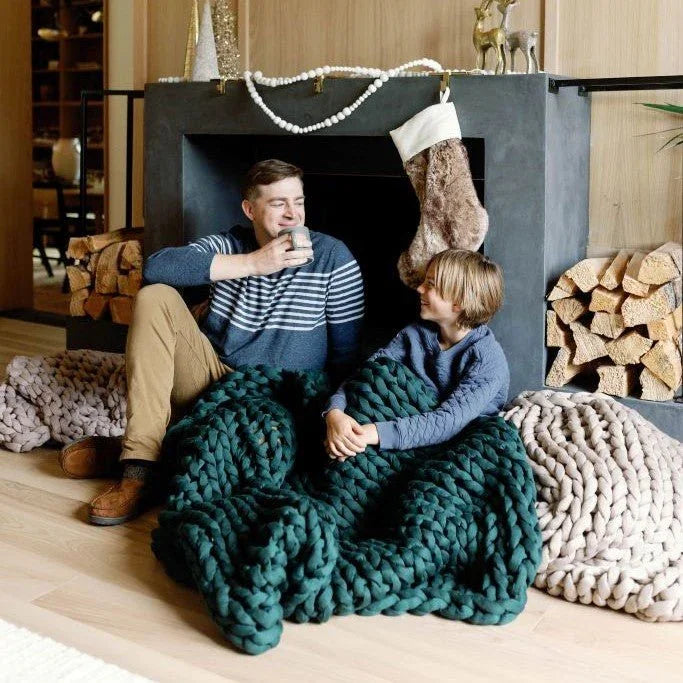 Father and son relaxing by fireplace under chunky knit weighted blankets, cozy living room setting