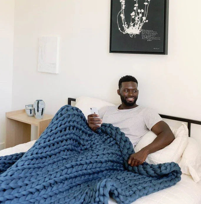 Man relaxing in bed with a chunky knit blue weighted blanket, holding a phone