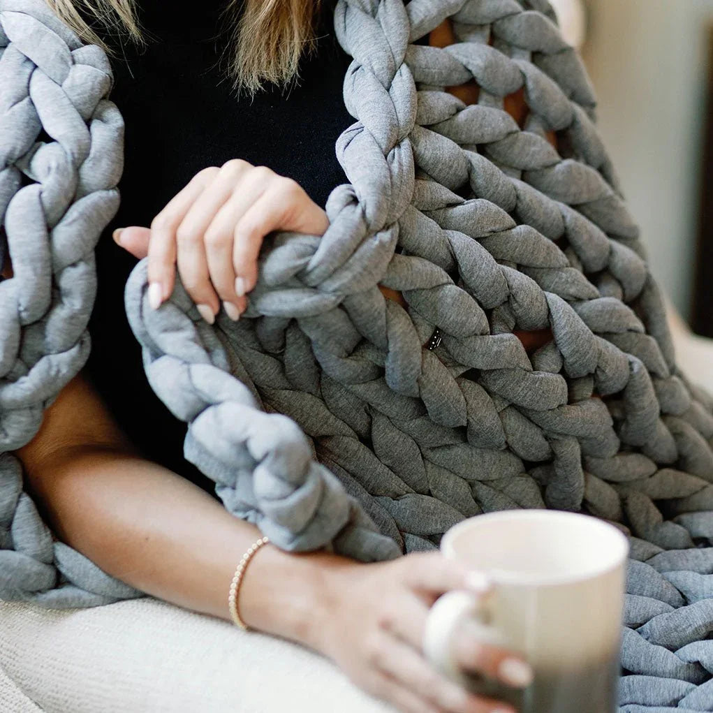 Person relaxing with a chunky knit grey weighted blanket and holding a mug