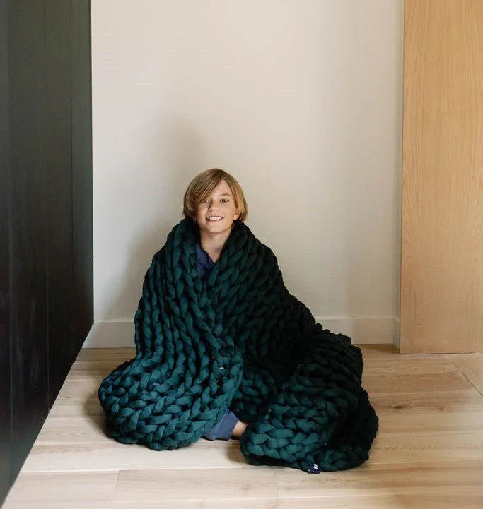 Smiling child wrapped in a chunky forest green sensory weighted blanket on wood floor