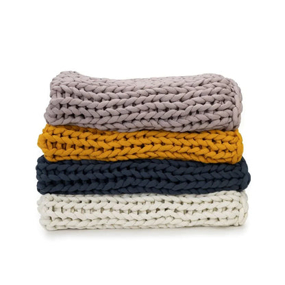 Stack of four chunky knit blankets in rose, marigold, blue, and cream colors