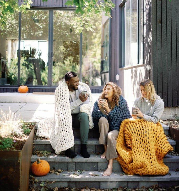 Three adults sitting on steps outdoors, wrapped in chunky knit blankets, holding mugs, with pumpkins nearby.