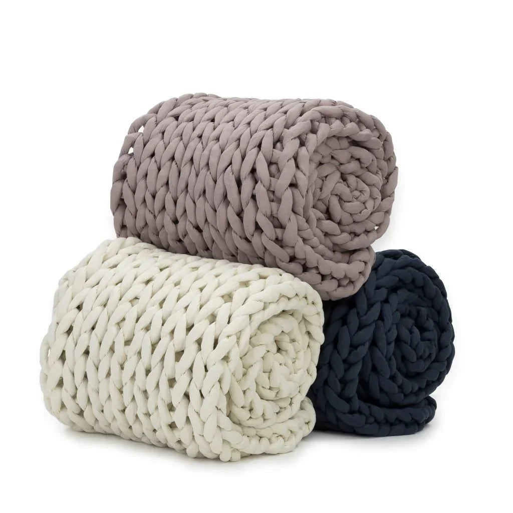 Three chunky knit weighted blankets in cream, blue, and taupe, rolled and stacked.