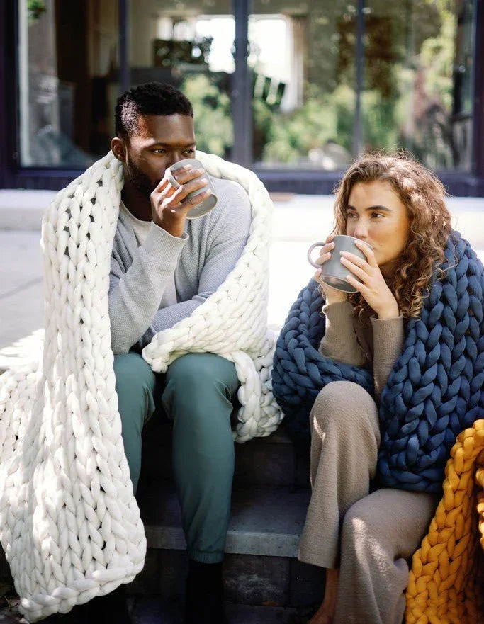 Two adults relaxing outdoors with mugs, wrapped in chunky knit weighted blankets