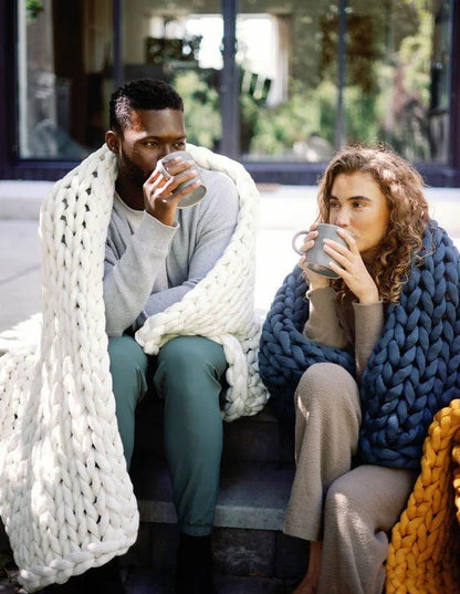 Two adults relaxing outdoors with mugs, wrapped in chunky knit weighted blankets