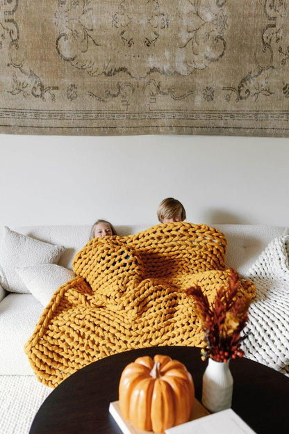 Two children cozy on sofa under chunky knit yellow blanket, fall decor in foreground