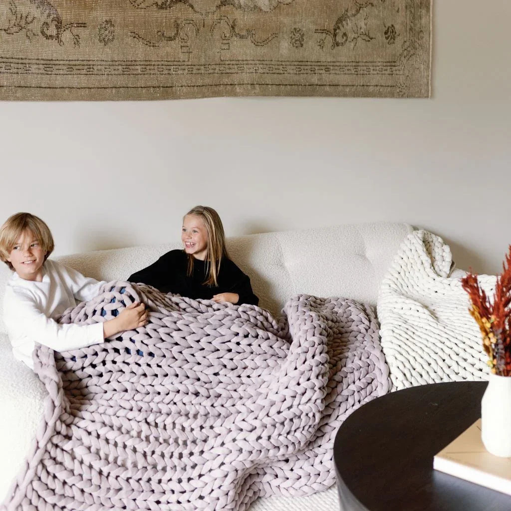 Two children relaxing on a white sofa under a chunky knit dusty rose blanket in a cozy living room