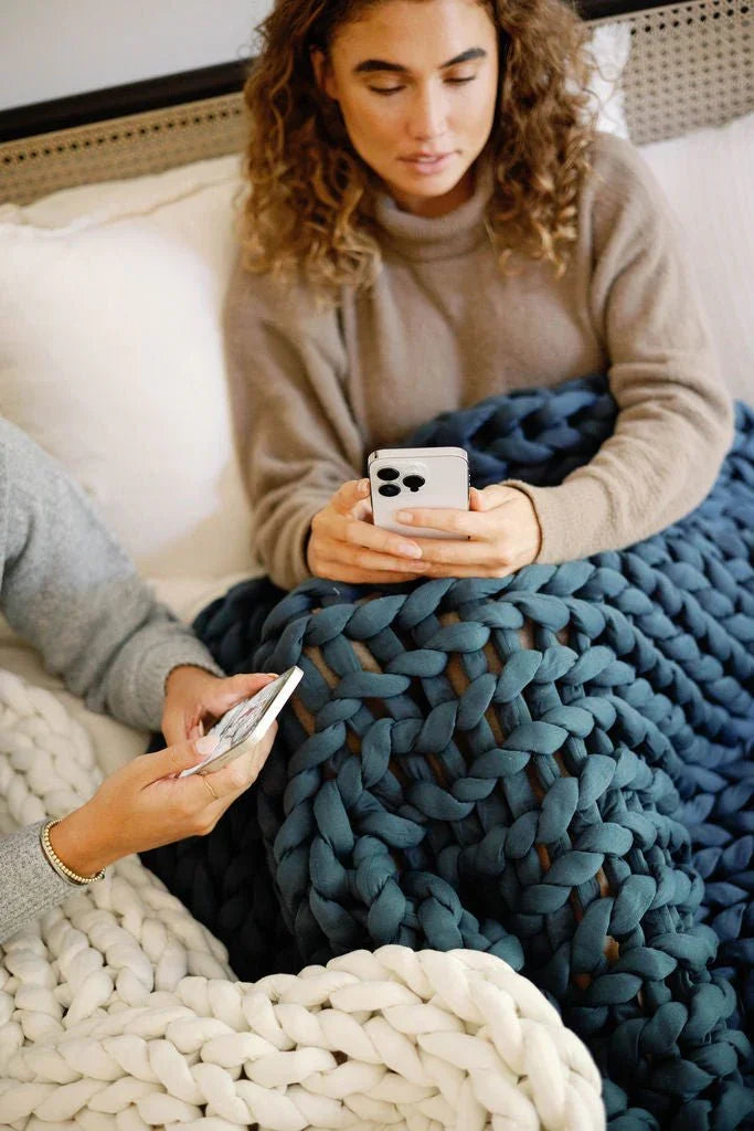 Two people relaxing under chunky knit sensory blankets, using smartphones in bed