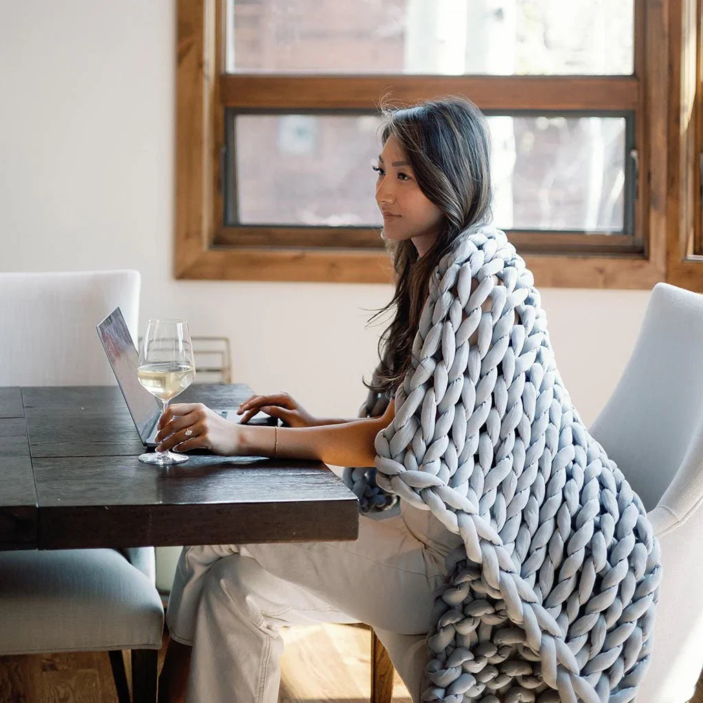 Woman working on laptop at dining table, draped in chunky knit sensory blanket, bright room