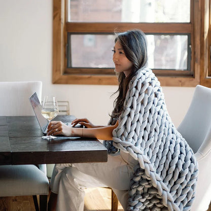 Woman working on laptop at dining table, draped in chunky knit sensory blanket, bright room