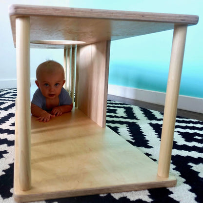 Baby crawling through wooden sensory play tunnel on patterned rug