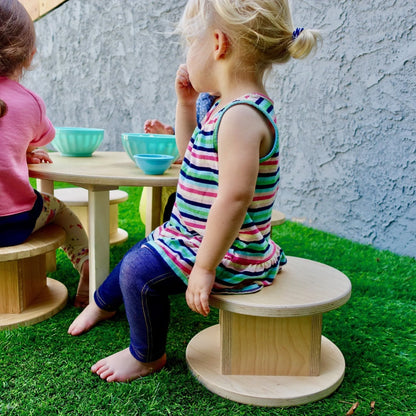 Young children sitting on round wooden stools at a low table outdoors on artificial grass