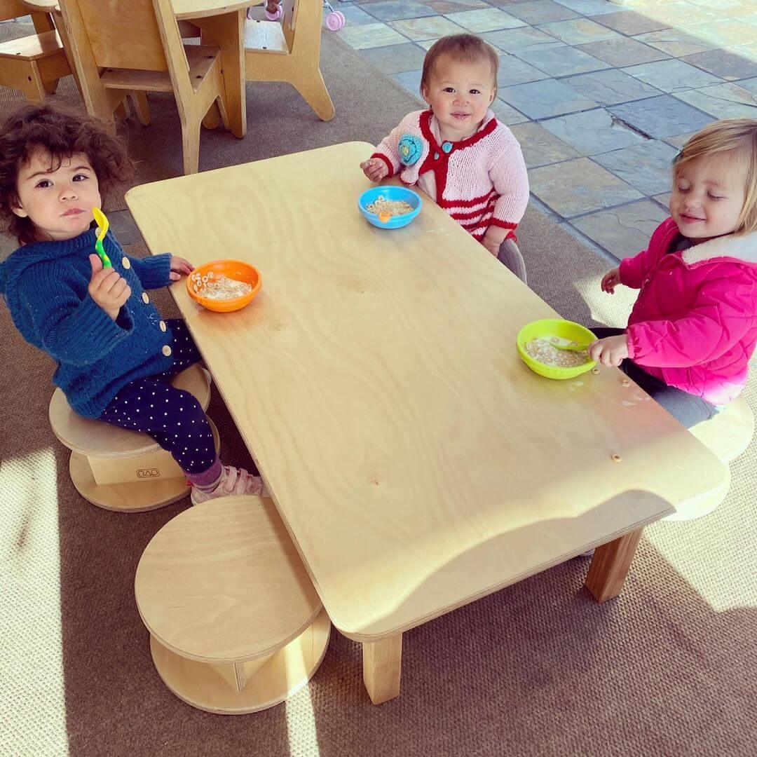 Three toddlers eating cereal at a low sensory-friendly wooden table with stools
