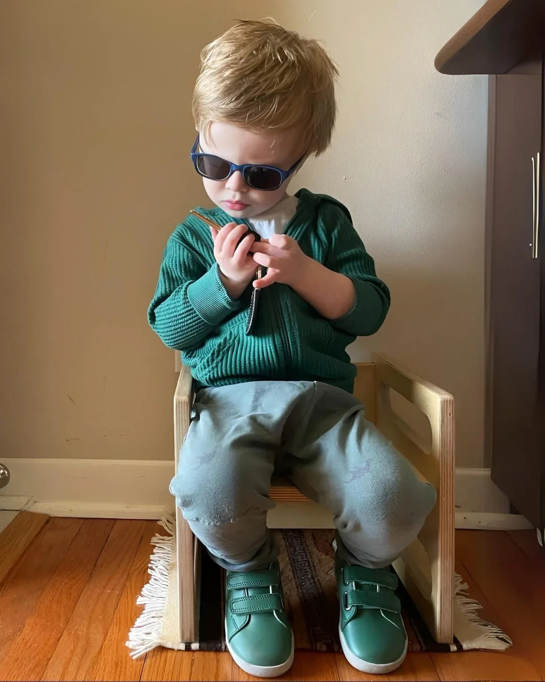 Toddler in sunglasses sits on wooden sensory cube chair, wearing green shoes and sweater