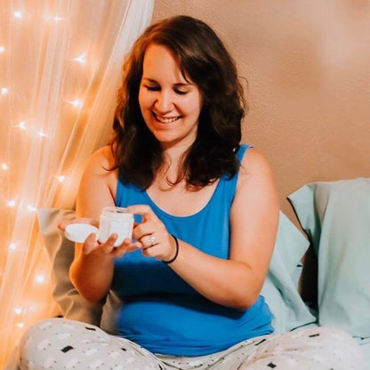 Smiling woman in blue pajamas sitting on bed holding a jar of cream, cozy bedroom setting.