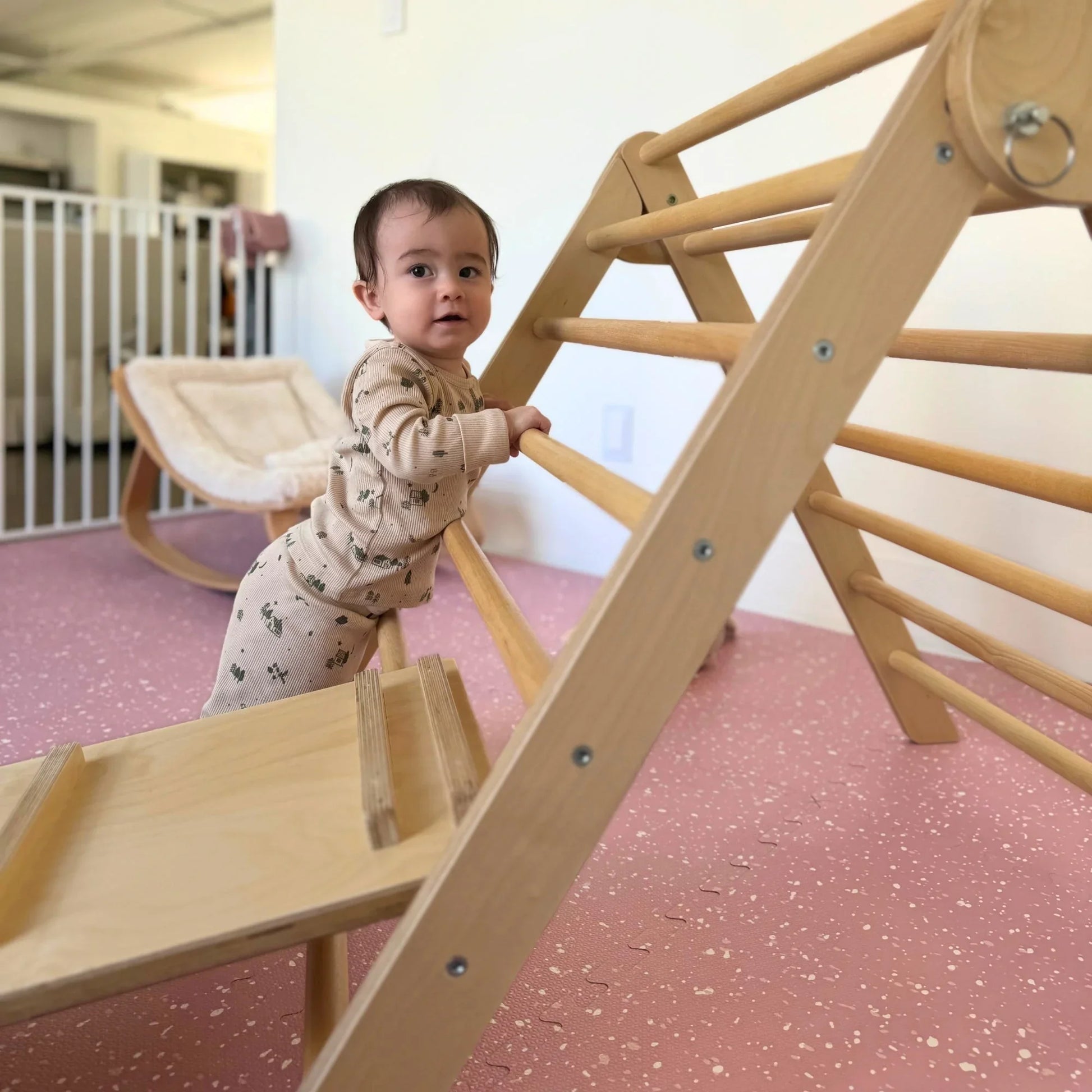 Baby in sensory pajamas playing on wooden climbing triangle in a safe indoor playroom