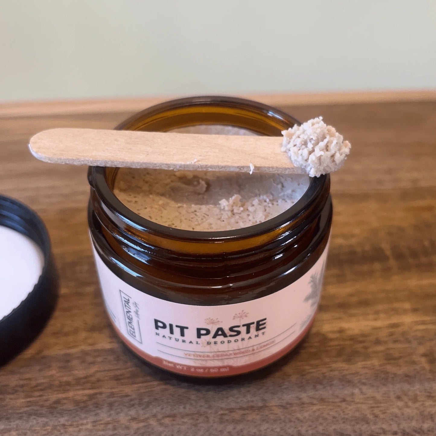 Open jar of all-natural pit paste deodorant with wooden applicator on wooden surface
