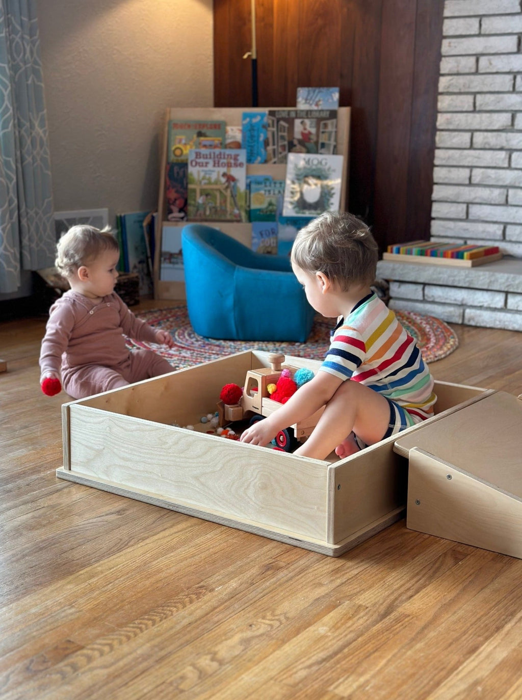 Two children playing with toys in a wooden box in a room with books and a blue chair.