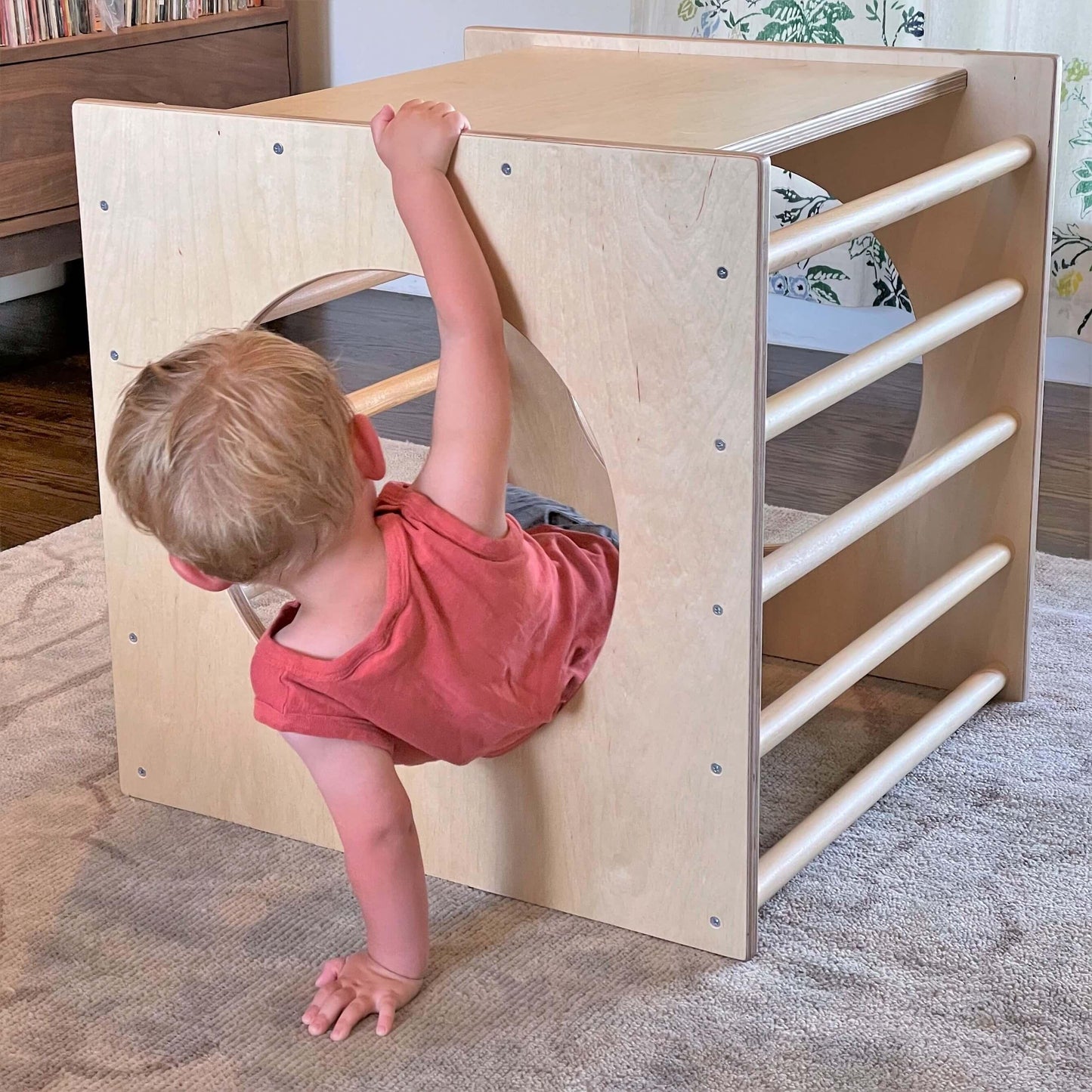 Toddler climbing out of a wooden sensory play cube on a rug in a cozy room.