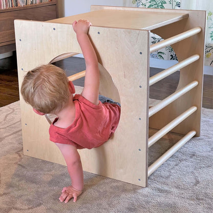 Toddler climbing out of a wooden sensory play cube on a rug in a cozy room.
