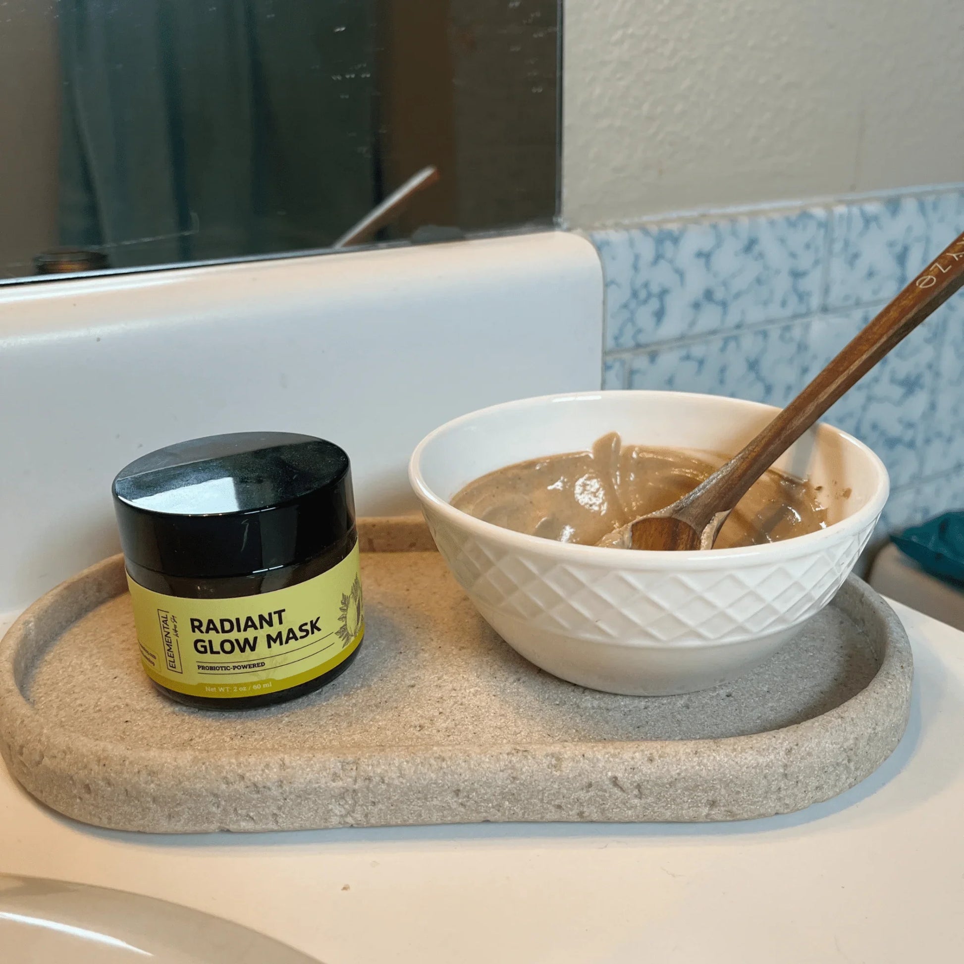 Radiant Glow Mask jar next to a white bowl with face mask mixture and wooden spoon on a bathroom tray