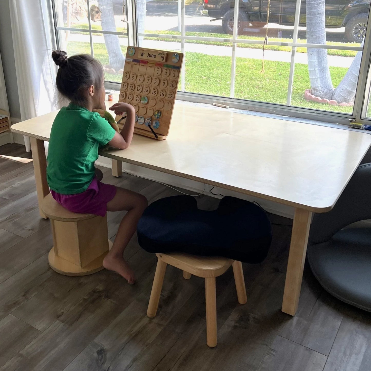 Child at wooden rectangular homeschool table with calendar, bright room, sensory seating