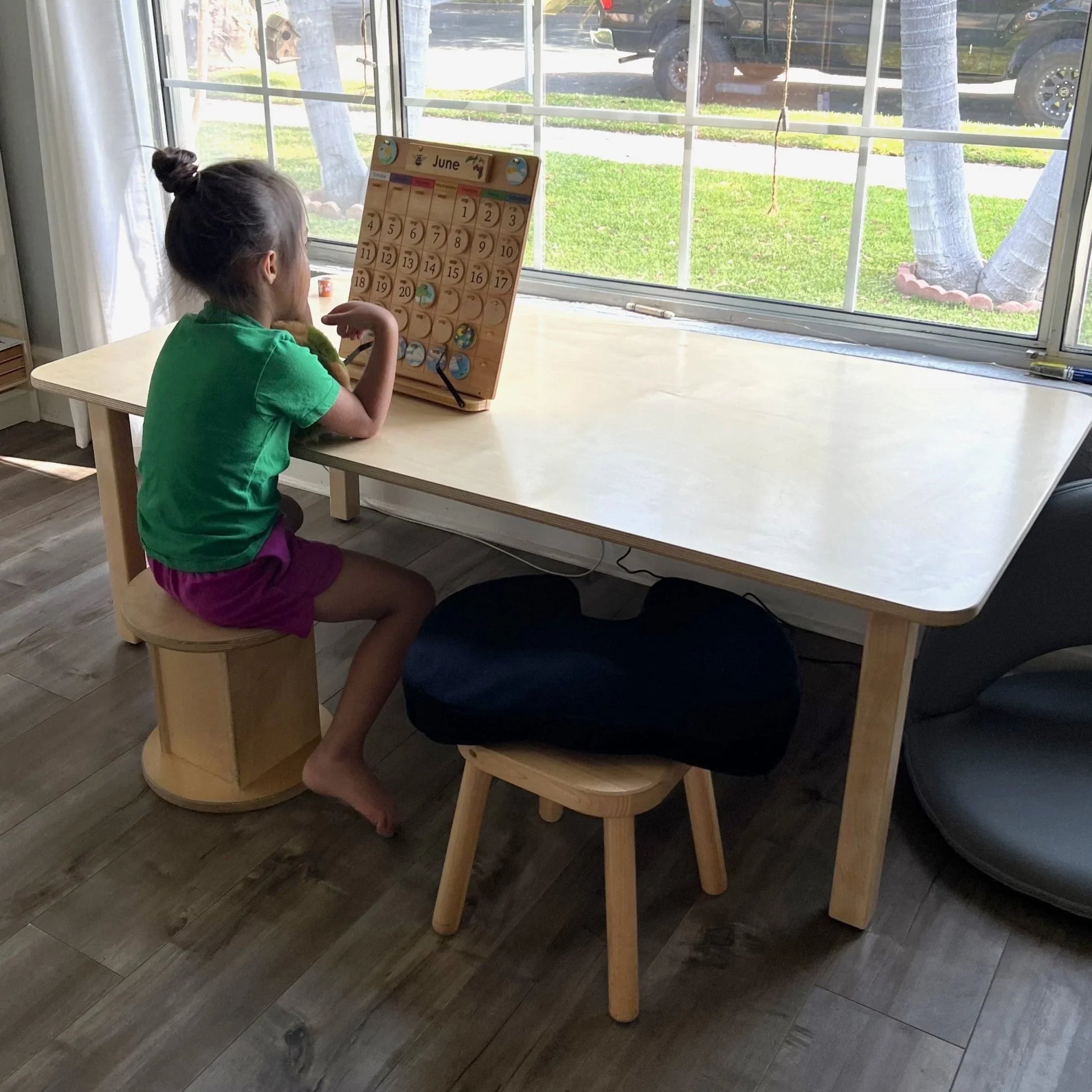 Child at wooden rectangular homeschool table with calendar, bright room, sensory seating