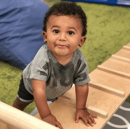 Toddler climbing on a wooden reversible climbing ramp in a sensory play area