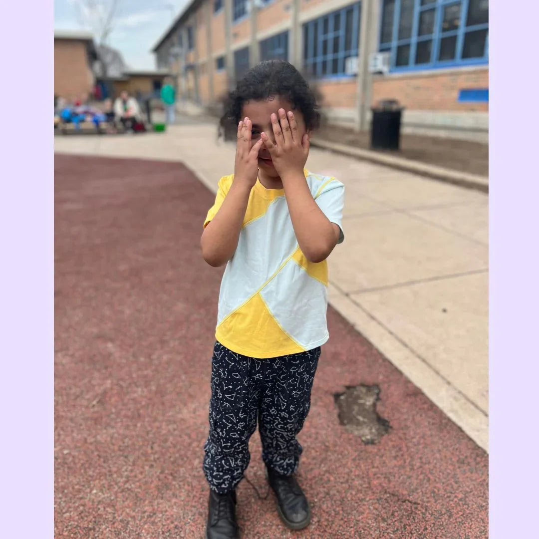 Child in sensory-friendly shirt and patterned pants covering face outside school building
