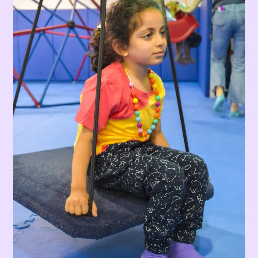 Child wearing sensory-friendly pants with playful patterns, sitting on an indoor swing
