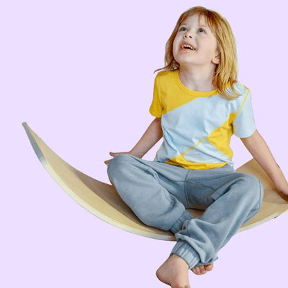 Smiling child in sensory-friendly joggers sitting on a wooden balance board against a pastel background.