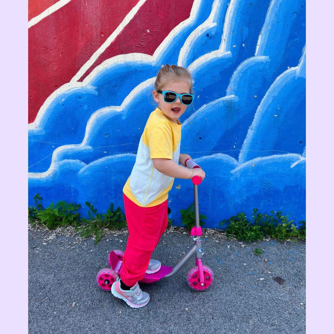 Young child in sensory-friendly clothing rides a pink scooter in front of a blue mural