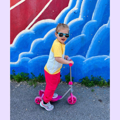 Young child in sensory-friendly clothing rides a pink scooter in front of a blue mural
