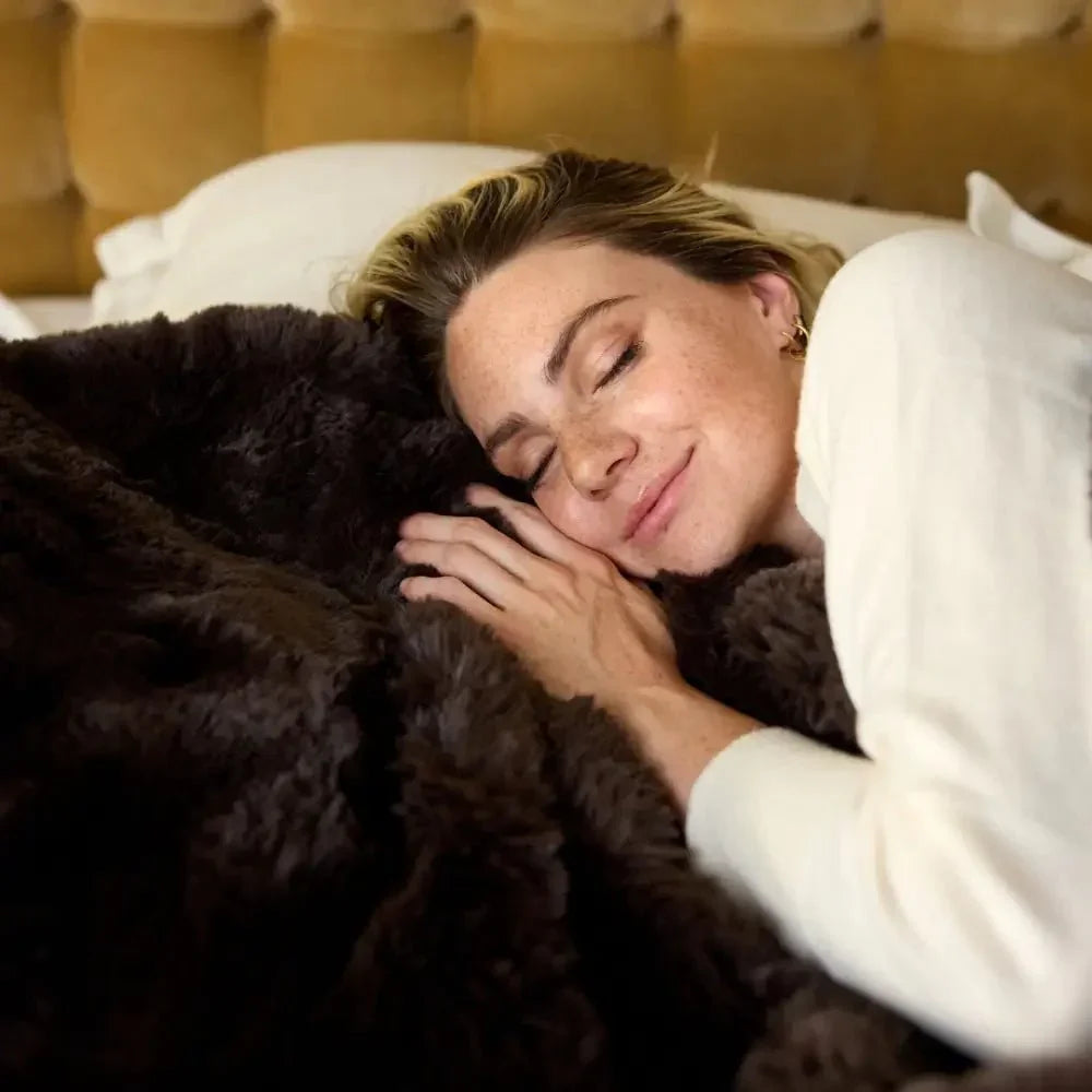 Woman smiling and resting under a soft, mocha weighted blanket on a bed