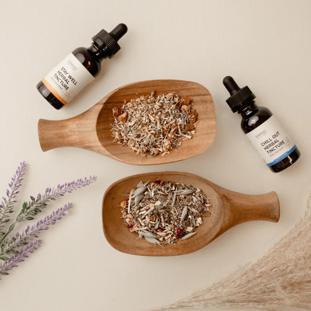 Two wooden trays with loose dried herbs and two bottles of herbal tincture on a neutral background