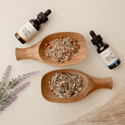 Two wooden trays with loose dried herbs and two bottles of herbal tincture on a neutral background