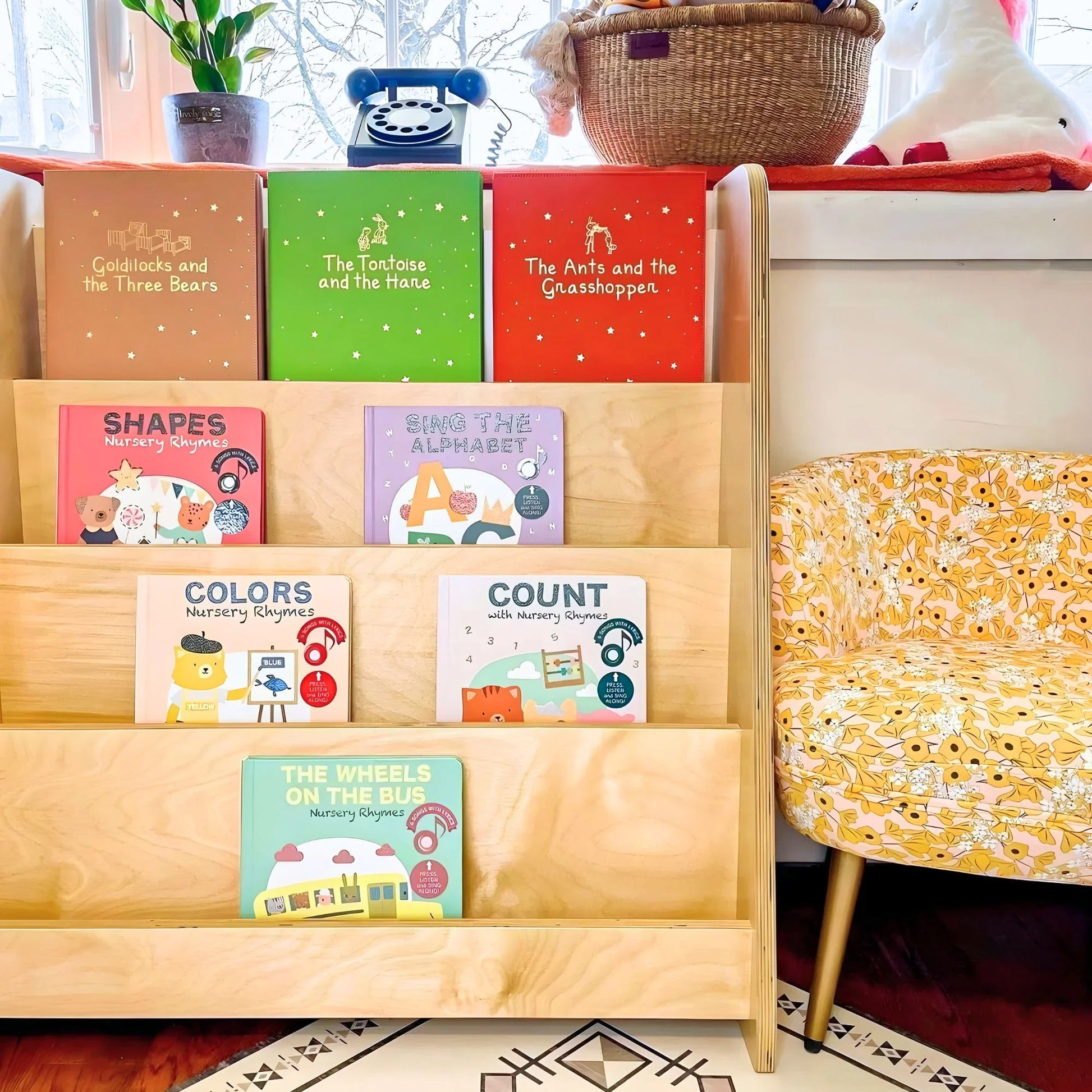 Montessori bookshelf with colorful children's books in a bright living room, next to a floral chair.