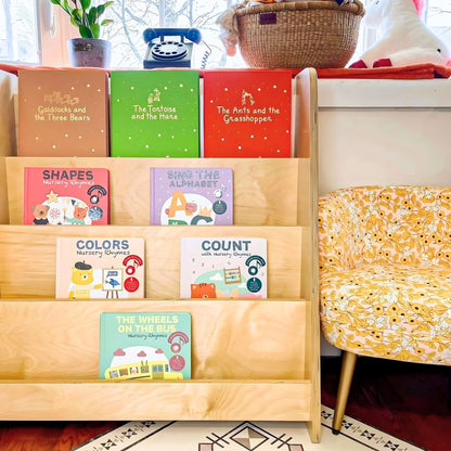 Montessori bookshelf with colorful children's books in a bright living room, next to a floral chair.