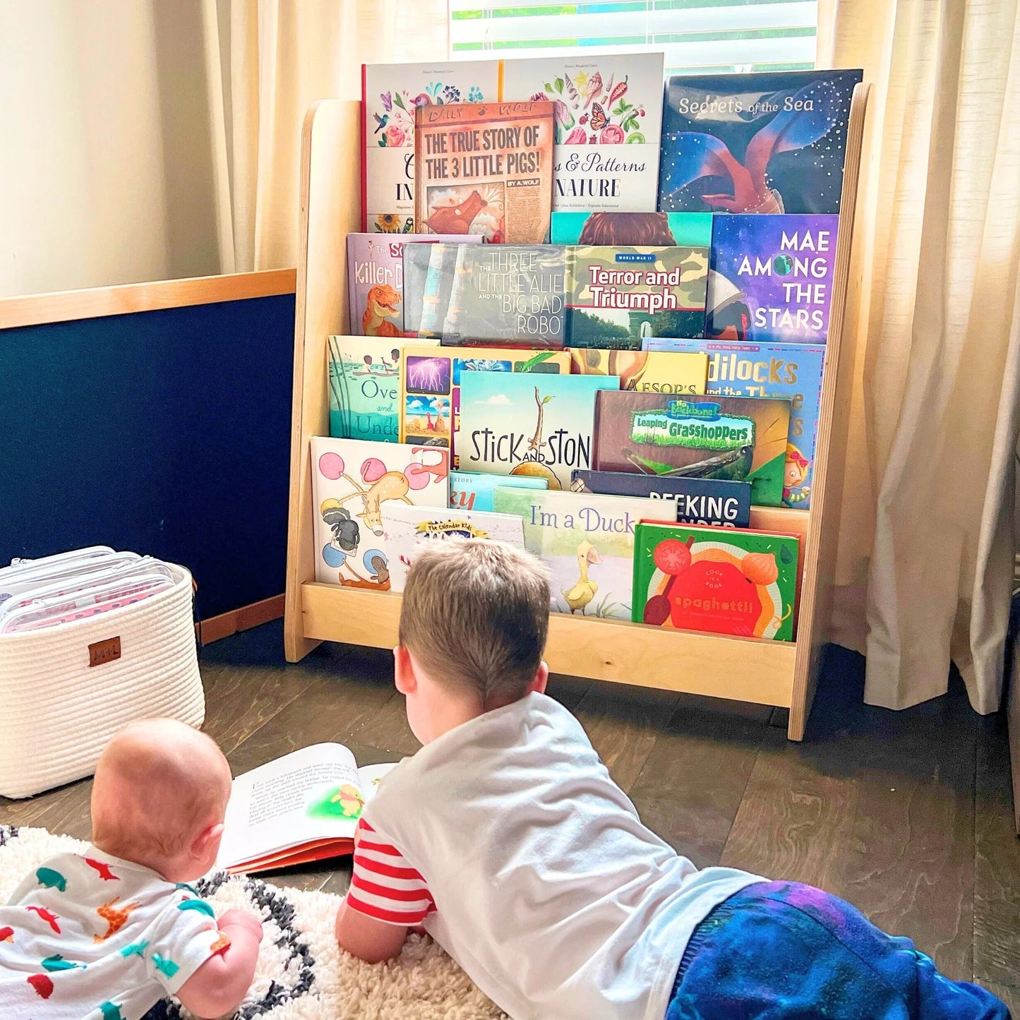 Two children reading on floor in front of a Montessori bookshelf filled with picture books