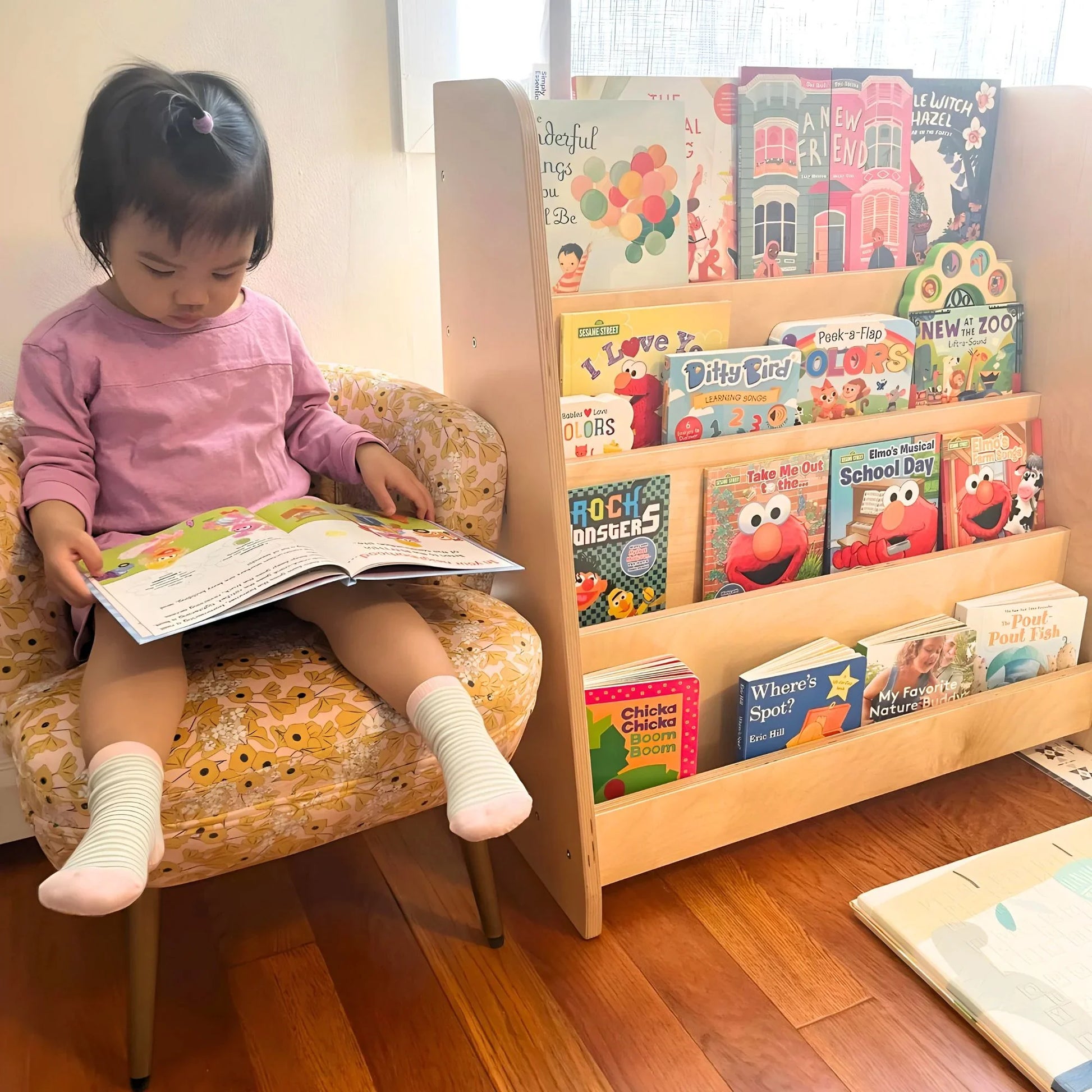 Toddler reading on chair beside Montessori bookshelf with colorful children’s books