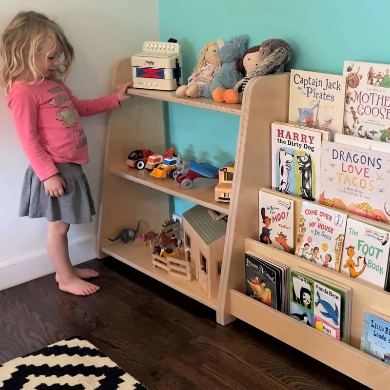 Toddler standing by a Montessori bookshelf filled with picture books, toys, and plush animals.