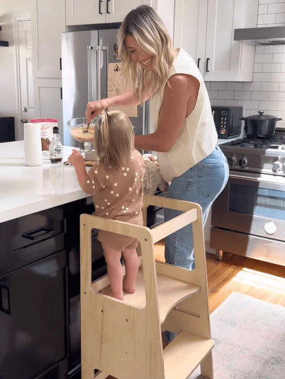 Mother and toddler using a wooden learning tower in a modern kitchen, baking together