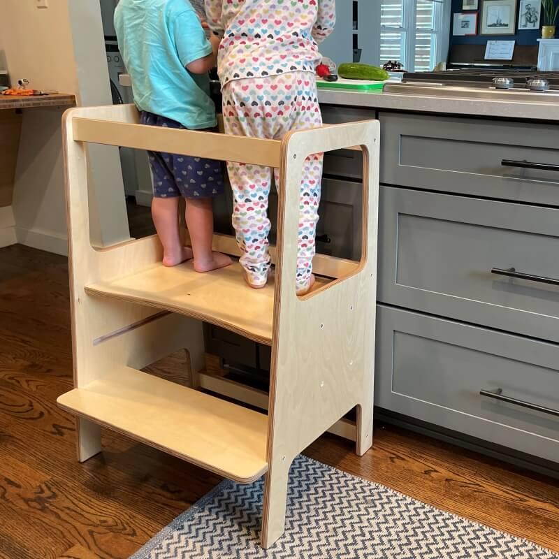 Two young children stand on a wooden learning tower in a kitchen, engaging at the counter.
